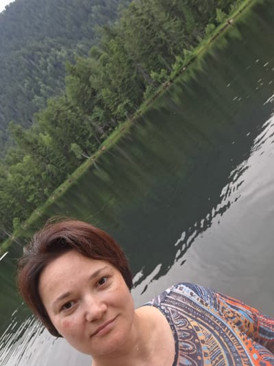 An outdoor, eye-level, selfie shows a woman with short, brown hair and fair skin with freckles, looking at the camera. She is wearing a colorful, patterned top and is positioned in the foreground with a lake and a forested hillside in the background. The lake's surface reflects the green trees on the hillside. The scene is likely in or near Vydrino, Russia, given the natural landscape.