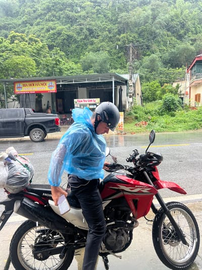 In Sơn Dương Commune, Vietnam, a person wearing a blue plastic poncho and a black helmet is standing next to a red and black Honda motorcycle on a wet, paved road. The person is holding a white smartphone in their right hand. The motorcycle is equipped with a black exhaust pipe and a spare tire mounted on the rear. A black pickup truck is parked behind the motorcycle. In the background, lush green hills covered in trees are visible under a rainy sky. A building with a red and yellow sign that reads "BIA HOI HANOI ANH EM QUAN" is situated behind the pickup truck. Another smaller sign with Vietnamese text is also visible. The environment suggests a rural or suburban setting during inclement weather.