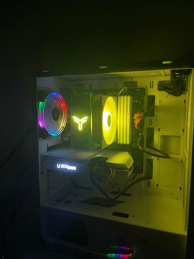 The image displays the illuminated interior of a desktop computer tower, located in the city of Ribnovo, Bulgaria. The PC case appears to be predominantly white or light-colored, showing some dust accumulation on internal surfaces and component shrouds.
Within the case, several components are clearly visible and active, emitting light. On the left side, a circular cooling fan exhibits multi-color RGB illumination, cycling through various hues. In the central upper section, a large CPU air cooler is installed, featuring a prominent, yellow-glowing stylized 'Y' or 'T' logo on its heat sink shroud. To the right of this CPU cooler, a cooling fan and several RAM modules are brightly illuminated with a consistent yellow light. A dark-colored motherboard serves as the foundation for these components.
At the bottom-center, a white-shrouded graphics card is horizontally mounted, with the brand "ZOTAC GAMING" illuminated in a pale white or light blue. The side of the card also displays "RTX". Power cables, including a bundled set with red/orange sleeving, are connected to components and routed within the case. Empty drive bays or mounting points are visible on the far right side of the case. The overall environment within the PC case is brightly lit by the internal component lighting, creating a high contrast against the dark, unlit background of the surrounding room.