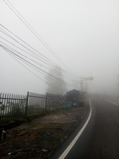 The image displays a roadside scene in Siruppiddy, Sri Lanka, heavily obscured by dense fog. An asphalt road, marked with a solid white line on its right edge, curves gently to the right, receding into the fog. To the left of the road, a dark-colored, vertically slatted fence runs parallel to the road, beyond which is indistinct vegetation. Adjacent to the fence and slightly elevated from the road surface stands a small, makeshift stall or shack with a dark-colored tarp roof. In front of this stall, approximately eight to ten light green plastic chairs are arranged. Numerous black utility lines crisscross the upper portion of the frame, originating from the left and extending towards the right, disappearing into the pervasive fog. Further down the road, a utility pole is visible, supporting at least two yellow diamond-shaped warning signs, all partially obscured by the fog. No individuals are present in the scene.