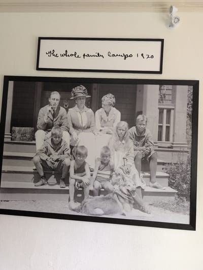 The image shows a framed black and white photograph of a family and a dog posed on the steps of a house. Above the photograph is a framed document with handwritten text that reads "the whole family Campobello 1920". The family consists of a man, a woman, and four children, all dressed in clothing appropriate for the early 20th century. The man is seated on the top step, smoking a cigarette and looking to his left. The woman is seated next to him, wearing a hat and looking forward. Two boys are seated on the steps below them, one in the center and one to the right. A girl with long hair is seated between the two boys. A dog is lying on the ground in front of the steps. The house appears to be a large, wooden structure with a porch and windows visible in the background. The setting appears to be outdoors, on a bright day, with foliage visible around the house. The photograph is framed with a black border and hung on a white wall, with a white emergency light fixture mounted on the wall above and to the right of the framed text.