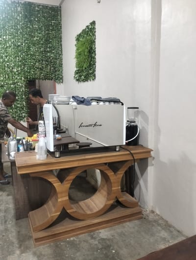 This media file captures a scene inside a coffee shop in Tingkeummanyang, Indonesia. Two men are visible, appearing to be employees or owners working behind a counter. One man, wearing a camouflage shirt, is on the left, seemingly in the process of preparing something. The other man, on the right and wearing a dark t-shirt, is looking towards the espresso machine.
The focal point of the image is a professional-grade white espresso machine, branded with "ferratti ferro" in elegant script. It is situated on a wooden counter with a distinctive, decorative base featuring large, curved wooden elements. To the right of the espresso machine, a dark-colored coffee grinder with a transparent hopper is visible. On the counter, there are stacks of white plates, cups, and various beverage preparation tools. A plastic bottle with a white label is also present. The shop's interior is decorated with artificial green foliage, hanging from a wire mesh on the left wall and as a rectangular accent piece on the right wall. The walls are painted a light, neutral color, and the floor appears to be concrete.
The lighting suggests it could be evening or an indoor setting with artificial lights, as there are distinct shadows cast on the wall. The overall atmosphere seems to be one of quiet operation, possibly during a lull in customer activity. The scene is factual and devoid of overt emotional expression, focusing on the operational aspects of the coffee shop.