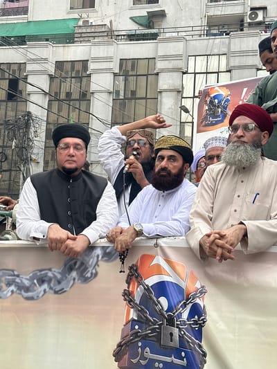 This media file depicts a group of men gathered outdoors, likely for a protest or religious gathering. The subjects are predominantly men, appearing to be of South Asian descent, dressed in traditional attire. Several individuals are wearing turbans, including a prominent figure with a maroon turban and a long grey beard, who is looking directly at the camera with glasses. Another man, wearing a white shirt and a black vest, is also in the foreground. The background features a multi-story building with numerous windows, suggesting an urban setting. Overhead, a dense network of electrical wires is visible, characteristic of many urban areas in Pakistan. The overall atmosphere seems serious, with the expressions of the individuals suggesting a focused or perhaps concerned mood.
A key object in the scene is a large banner or poster held up by some of the individuals. This banner features a graphic of a lock and chains, symbolizing confinement or suppression, with some text visible in Arabic script on the banner's graphic. The time of day appears to be daytime, with soft lighting that doesn't cast harsh shadows. The weather seems to be overcast or perhaps it is late afternoon, as the light is diffused. The image is captured with a slightly upward angle, emphasizing the subjects and the banner. The presence of the building and the overhead wires strongly points to a location within a Pakistani city, with Karachi being specified as a potential location context. The focus of the image is on the men and the symbolic imagery on the banner, indicating a message of protest or solidarity.