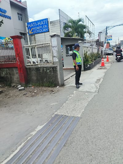 A man in a neon vest stands on a sidewalk near a "Hati-Hati Komplek Sekolah" sign, which translates to "Caution School Complex." He's wearing a navy hat and black pants. The sidewalk has tactile paving for the visually impaired. Behind him is a building and a fence with a guard booth. Orange traffic cones line the sidewalk further down the road. A motorcycle and car are visible in the distance. The sky is overcast.
