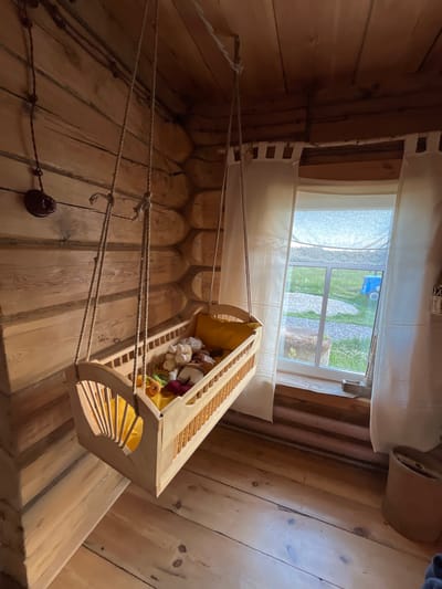 This image shows an interior scene of a room with wooden walls and floors. Suspended from the ceiling by ropes is a wooden cradle filled with stuffed animals. The cradle has a yellow interior lining and appears to be a child's toy or bed. Through a window in the background, a green, grassy area with some gravel paths and what appears to be a blue toy truck is visible, suggesting an outdoor environment. The window is covered by sheer white curtains. The lighting suggests it might be daytime. The room has a rustic aesthetic, with the wooden logs forming the walls being particularly prominent. There is a dark, circular object attached to the wall near the bottom left, which could be a light fixture or a similar mounting. The perspective of the photo is from a low angle, looking upwards towards the cradle and the window. The overall impression is one of a cozy, perhaps child-friendly, space within a wooden structure.