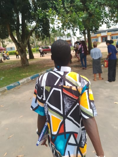 The image depicts an outdoor scene in Alakahia, Nigeria, specifically on what appears to be the campus of the University of Port Harcourt (UNIPORT), as indicated by a visible lanyard. In the foreground, a dark-skinned male individual, viewed from behind, walks rightward on a paved path. He wears a short-sleeved button-up shirt with a distinctive geometric pattern in black, white, yellow, orange, green, blue, and light grey. His left hand holds a blue and white lanyard or strap bearing the text "I LOVE UNIPORT." To his left, a curb with blue and white painted stripes runs alongside the path.
In the midground, a diverse group of people is visible. On the far left, several individuals are gathered under a red canopy. A red sedan is parked or stationary on a paved area, with multiple people standing near it. Further right, a woman with long dark hair wears a light pink shirt and blue jeans, walking away. Adjacent to her, a woman in a light blue shirt and a dark skirt stands facing away from the camera, wearing sandals. To the far right, another woman, dressed in a dark blue shirt and dark trousers, stands with her back to the viewer, holding a red object, possibly a bag or container, in her left hand.
The environment is characterized by lush green foliage from numerous mature trees that line the area. In the background, several multi-story buildings with light-colored facades and architectural accents in yellow and blue are discernible, consistent with institutional architecture. The ground consists of paved surfaces, dirt patches, and areas of grass. The scene is illuminated by natural daylight.