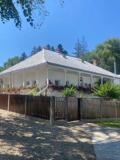 The image depicts a white, single-story house with a wide, covered porch supported by white columns. The porch is decorated with flower boxes filled with red and pink flowers. A wooden fence runs along the front of the property, with a gate visible. A gravel path leads to the gate, and to the right of the path is a paved walkway. The house has a metal roof and two chimneys. Trees and foliage are visible behind the house and along the fence line. The sky is clear and blue. The scene is located in the city of Filioara, Romania. There are no people or animals visible in the image.