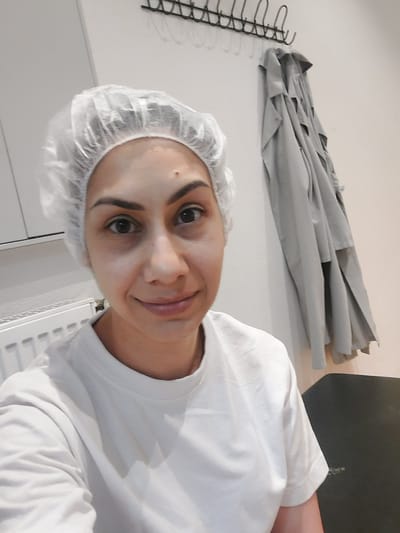 The image shows a person with fair skin wearing a white t-shirt and a disposable white hairnet. The person is likely indoors based on the clothing and hairnet, potentially in a sterile or sanitary environment like a kitchen, laboratory, or medical facility. Behind the person is a light-colored wall with a radiator and white cabinet. There is a light gray garment, possibly a coat or lab coat, hanging on a metal rack with hooks attached to the wall. The photo is a close-up, selfie-style shot. There is no information to determine the location in Altdorf, Germany.