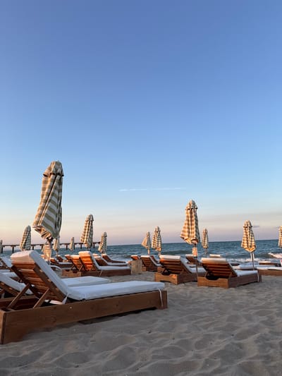 The image shows a beach scene at Shkorpilovtsi, Bulgaria. The foreground is dominated by sand, with several rows of lounge chairs and closed umbrellas lined up. The chairs are made of wood with white cushions, and the umbrellas have a striped pattern of green and beige. The sea is visible in the background, with gentle waves rolling towards the shore. The sky is a clear, light blue, with a faint contrail from an airplane visible. The lighting suggests it is either early morning or late afternoon, with a soft, warm glow. There are no people visible in the image, giving it a peaceful and serene atmosphere. The scene evokes a sense of tranquility and relaxation, typical of a beach resort.