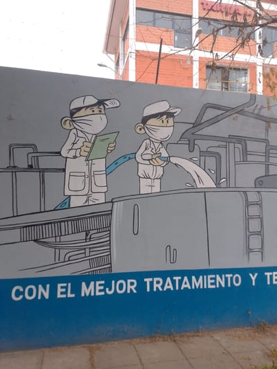 A close-up shot of a painted wall in Municipio Tarija, Bolivia. The painting depicts two cartoon figures wearing caps, white lab coats and protective masks, standing on elevated platforms or tanks. The figure on the left is holding a green clipboard. The figure on the right is pouring a stream of liquid from a container into a large tank. The background features industrial equipment and tanks rendered in a cartoon style. Below the cartoon image, in white text on a blue stripe, is a phrase in Spanish "CON EL MEJOR TRATAMIENTO Y TE". Behind the wall is a multi-story building with orange brick facade, and some trees.