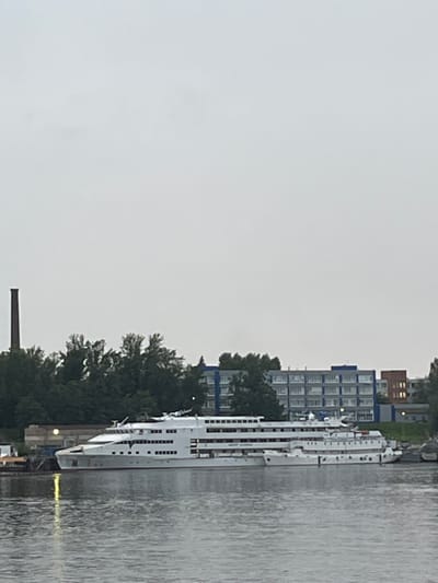 The image shows a large, white cruise ship docked in a body of water. The ship has multiple decks with numerous windows and lifeboats visible. Behind the ship, there is a line of lush green trees. Further in the background, a multi-story building complex with blue trim and rows of windows is visible. To the left of the trees, a tall, dark brick chimney stands against a cloudy, overcast sky. The water in the foreground reflects the overcast sky and the ship. The scene depicts a quiet, possibly industrial, waterfront area in the city of Imeni Sverdlova, Russia.