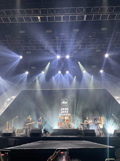 The image depicts a live music performance at the 2023 Java Jazz Festival, located in Salembaran, Indonesia. A band is on stage, lit by numerous spotlights, in front of a large screen displaying the festival logo. The band includes a drummer, keyboard players, a saxophonist, and a bassist. A metal truss structure supporting the lighting is visible. A person can be seen in the foreground, holding a mobile phone, likely recording the performance.