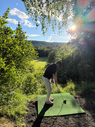 In Lommedalen, Norway, on a bright, sunny day, a person is standing on a green golf tee mat, holding a golf club in preparation to swing. The individual is wearing a black t-shirt, light-colored pants, and a baseball cap. The tee mat is situated on dark gravel, with patches of green grass and foliage surrounding it. In the background, a lush green golf course is visible, with trees lining the fairway and a distant hillside dotted with buildings. The sky is a clear blue with scattered white clouds, and the sun shines brightly through the trees, casting a lens flare onto the image.