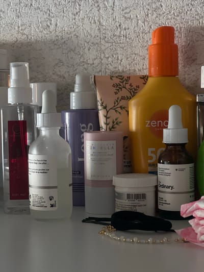 This image shows a shelf in a residential bathroom, likely in Genemuiden, Netherlands. On the shelf are various skincare and personal care products. From left to right, there is a clear spray bottle labeled "BODY MIST" with a red label, a white dropper bottle, a purple bottle, a cylindrical pink container, and a large yellow bottle with an orange cap, labeled "zendium sunscreen". There is also a small brown dropper bottle labeled "The Ordinary". Several hair accessories, including bobby pins and a beaded bracelet, are visible in the foreground. The background features a textured, light-colored wall. The lighting is soft, suggesting ambient indoor light. There are no people present in the image.