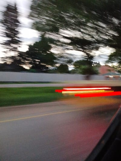 A blurry, motion-filled scene presents a blurred view of trees and sky on the left. A solid, neutral-toned barrier or structure runs vertically down the center. To the right, a blurred green field transitions into a smooth, gray surface, perhaps a road or building. Vertical red and yellow streaks suggest lights or reflections, further emphasizing the sense of speed. The angle indicates it might be a shot through a car window.