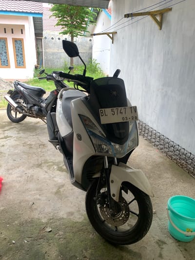 The image depicts two motorcycles parked in an outdoor space, likely a residential area in Blangbladeh, Indonesia. The primary motorcycle, a white Yamaha, is prominently positioned towards the foreground, showcasing its front wheel, fender, and a license plate bearing the registration "BL 5747 TV 03.29". Behind it, a second, black motorcycle is partially visible. The ground is concrete. To the right of the white motorcycle, a light green bucket sits on the concrete. In the background, a wall and a fence are visible. The corner of a house with a red tile roof and two windows is also observable. Above the motorcycles, a wooden structure with attached cords extends from the ceiling or roof. A tree is also visible in the background.