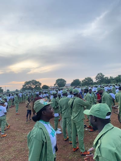 A large outdoor gathering of several hundred individuals, primarily young adults, is depicted on a reddish-brown dirt field in Kubi A, Nigeria. The majority wear a uniform consisting of light green long-sleeved shirts or jackets, green cargo pants, and green baseball caps, characteristic of the Nigerian Youth Service Corps (NYSC). Many are also wearing bright orange shoes or sandals.
In the left foreground, a woman in glasses, a green cap, and a green jacket over a white t-shirt with "NYSC" lettering, is smiling and looking towards the right. To her right, a man in a similar green uniform is partially visible from behind, looking towards the woman. A young child in a light-colored t-shirt and dark pants stands among the uniformed individuals in the mid-ground.
The crowd is densely packed across the field, with individuals standing and conversing. Several people are observed holding or interacting with mobile phones; one man in the mid-ground holds a phone to his ear. A bicycle wheel is partially visible on the far right. The background features a tree-lined horizon and distant, low-lying structures. The sky above is largely overcast with grey clouds, exhibiting soft orange and yellow hues near the horizon, consistent with either dawn or dusk lighting.