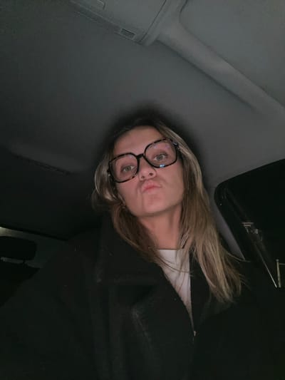 A woman wearing glasses and a black sweater is visible in a close-up shot. She is positioned in what appears to be a car, evidenced by the car ceiling and interior. The lighting is dim. The woman has blonde hair and is pouting her lips towards the camera. The image is angled, giving a tilted perspective of the subject and her surroundings.