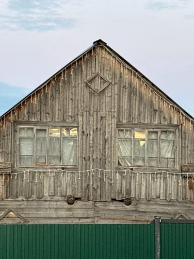 The image shows the facade of an old, wooden house with a gabled roof in the village of Kukui, Russia. The wooden siding is weathered and vertical. Two windows are visible on the upper floor, each with multiple panes. White string lights are draped across the facade below the windows. A diamond-shaped decorative element is centered above the windows, on the gable. Below the main wooden structure is a green metal fence. The sky above is overcast with hints of blue. There are no people or animals visible in the image.