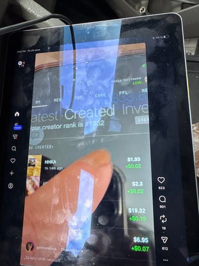 The image shows a tablet screen displaying a social media interface. The top left corner of the screen shows the time "3:42 PM" and the date "Пн 29 июня". The battery indicator in the top right corner shows 34% charged. The screen displays text such as "latest Created Inve" and "Your creator rank is #1332". There are also prices listed, such as "$1.93", "+$0.02", "$2.3", "+$0.02", "$19.32", "+$0.19", "$6.95", and "+$0.07". Below these are icons that appear to be for likes, comments, shares, and possibly more options, with counts such as 623, 901, 19, and 612. A profile picture and username "gellmundova" are visible, with the word "Подписаться" next to it. The text below the username reads "Za lajky rohlík nekoupíš". A person's finger is visible on the screen, obscuring part of the display. The tablet appears to be placed on a surface, and the background suggests it might be in a vehicle, given the presence of a dark, textured material that resembles a car's dashboard or interior. There are reflections on the tablet screen, indicating ambient light sources. The location is stated to be Mebane, United States of America.