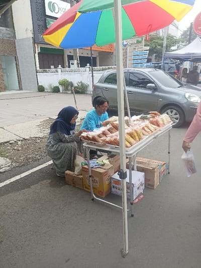 A street vendor is selling snacks from a folding table under an umbrella in Purwokerto, Indonesia. The vendor is a man in a blue shirt, wearing a black cap, and a woman in a hijab. The woman in the hijab is looking at the snacks on the table. The man is looking to the right, at the woman in the hijab. The table is full of snacks, packaged in clear plastic bags, and the vendor has several boxes of snacks stacked underneath the table. A silver car is parked behind the vendor, and a white line is visible on the road in front of the vendor. The vendor is located on the sidewalk next to the road. The vendor is standing next to a building with a sign that says "Restu Ibu". The vendor is selling the snacks to a woman in a pink shirt, holding a plastic bag full of snacks. The woman in pink is standing on the sidewalk. There are more people standing in the distance but they are blurred. The scene is set in a daytime. The overall atmosphere of the scene is of a busy, bustling street market.