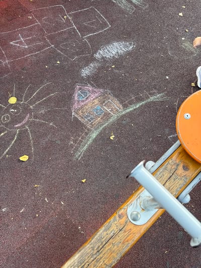 A child's drawing of a house and a sun is chalked on a dark reddish-brown rubberized playground surface. The drawing is located in the city of Vyazniki, Russia. A portion of an orange seesaw seat and its wooden and metal support structure are visible in the lower right quadrant of the image. The drawing of the house has a pink roof and blue windows. The sun is yellow with rays extending outwards. Several fallen leaves are scattered on the ground. A child's hand and shoe are partially visible in the upper right corner.