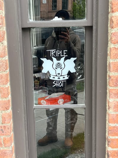 The media file displays a rectangular window, framed by a reddish-brown brick wall on the top and bottom, and dark grey painted wood on the sides and horizontally between the panes. The window itself is divided into multiple panes by dark grey wooden mullions.
A white sticker is affixed to the central panes of the window. The sticker features a stylized graphic of what appears to be an animal's head (possibly a bull or cow, with prominent horns and a patterned snout design) and the words "TRIPLE SHOT" written vertically to the left of the head, and "SHOT" (or possibly "SHOT" as part of a rotated "TRIPLE SHOT") vertically to the right.
Through the glass, various reflections are visible. A person is largely reflected across the left side of the window. This reflection shows an individual wearing a dark hoodie, camouflage-patterned pants, and brown boots. Their hand, with fingers slightly splayed, is visible on the left side, seemingly near the glass. The person's head is partially visible further left in the reflection, obscured by the window frame and appearing as if they are looking down or away. The reflection of their lower body (legs and boots) is clearly seen in the lower right pane of the window, appearing sideways.
Other reflections include a car wheel visible in the upper central pane and a general outdoor street scene with trees and what looks like a sidewalk or paved area, also appearing rotated in the reflections. Inside the window, behind the sticker, a red box with white text, including the word "Logan," is visible on the right side of the lower left pane.
The scene appears to be outdoors during daytime, with natural light illuminating the brick wall and creating the reflections. The setting is a building facade, in the City of Newburgh, USA.