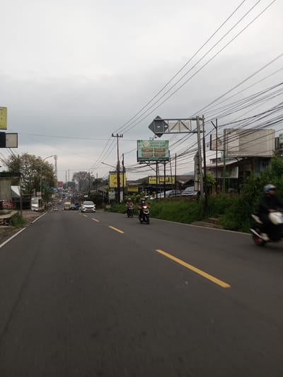 The image depicts an asphalt road in an urban or suburban area, likely in Asia based on the signage. The road has yellow center lines and leads into the distance where several cars are visible.  

Several power lines crisscross overhead. On the right side of the road, there's dense green vegetation and various advertising billboards. The billboards display text and graphics, with one prominently advertising "LAYAN SPA" in both English and Arabic. There is a restaurant called “MAMI NAUBA” .

Two motorcycles are on the road. One is in the foreground, and the rider is partially obscured. A second motorcycle with a rider is further up the road.

The weather appears overcast. The background includes buildings and trees. Based on the OCR content, it could be Taunggyi, Myanmar.