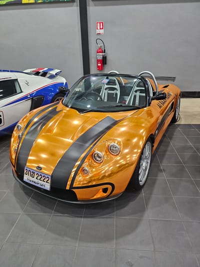 A bright orange convertible sports car with black racing stripes is parked on a dark tiled floor in what appears to be a showroom or garage. The car has a license plate that reads "3กฮ 2222" and "กรุงเทพมหานคร" (Bangkok). A white and blue sports car with a red, white, and blue spoiler is parked to the left of the orange car. In the background, a red fire extinguisher hangs on a grey wall. The scene is indoors, with overhead lighting reflecting off the glossy finish of the orange car. The overall impression is one of automotive display.