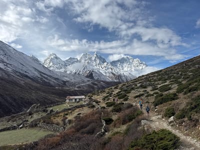 This wide-angle landscape photograph captures a majestic and rugged mountainous region in Khumjung, Nepal. Towering snow-capped peaks, characteristic of the Himalayas, dominate the background, their sharp ridges partially obscured by dramatic white clouds against a bright blue sky.
The midground and foreground showcase a steep, rocky hillside covered with sparse, dry vegetation, including low bushes and patches of light green grass. A winding dirt path, indicative of a trekking trail, traverses the slope from the lower right towards the middle left. Along this path, at least three hikers are visible, walking away from the viewer. One hiker wears a vibrant blue jacket and uses trekking poles, followed by another in a darker jacket carrying a backpack. A third, less distinct figure, is further up the trail.
On a terraced section of the hillside to the left of the path, a small, simple building with a light-colored exterior and a corrugated metal roof stands, suggesting a remote lodge or tea house. Numerous low, dry-stacked stone walls crisscross the slopes, segmenting the terrain around the building and alongside the trail.
The scene depicts an active trekking or hiking event under a cloudy but bright daytime sky, likely late morning or early afternoon. The weather appears cool, given the snow on the distant peaks. No visible text is present in the image. The overall atmosphere conveys the challenging yet awe-inspiring beauty of high-altitude mountain travel.