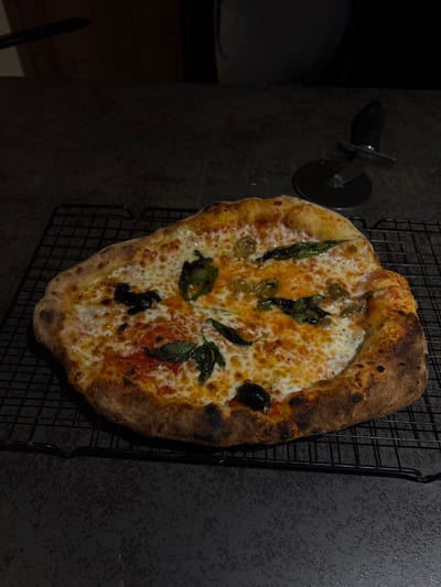 The image depicts a freshly baked pizza resting on a black wire cooling rack, positioned on a dark grey, speckled countertop in Ness Ziona, Israel. The pizza is roughly oval in shape with a puffed, golden-brown crust, showing some charring. It is topped with melted white cheese over a visible red tomato base. Scattered across the cheese are several dark green, slightly wilted basil leaves, light green/yellowish oval pieces (possibly green olives or pickled peppers), and a few dark, oval objects (possibly black olives). To the upper right of the pizza, a black-handled pizza wheel cutter with a silver blade is resting on the countertop. The scene is dimly lit, with the light concentrated on the pizza, and the background is dark and indistinct, suggesting an indoor kitchen setting. No individuals or active interactions are visible.