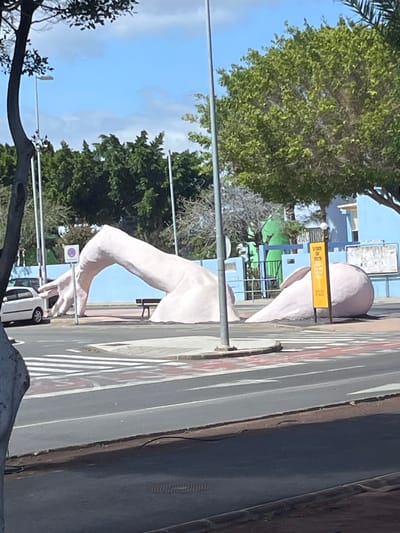 This image depicts an outdoor urban scene in La Unión, Spain, under clear daylight conditions. The central subject is a large, light pink-colored sculpture resembling a human arm and hand. The sculpture's hand rests near a pedestrian crossing on the left, while the arm arches over a paved area, with a substantial portion of the upper arm or shoulder appearing to emerge from or sink into the ground on the right.
The environment includes an asphalt road in the foreground and mid-ground, featuring white and red painted lines and a zebra-striped pedestrian crossing. A white two-door compact car is parked on the left side of the road.
Various objects are present: a dark brown slatted bench is partially visible behind the sculpture's arm, and a circular red and white "no stopping/parking" sign is mounted on a pole near the hand. To the right of the sculpture, a yellow vertical sign featuring an orange ribbon graphic is visible. A light blue building with a blue metal fence in front of it stands in the background, along with a white billboard or notice board displaying indistinct content. A tall, abstract green structure is situated behind the blue fence.
Green leafy trees are prominent on both sides of the frame and in the background, accompanied by some bare-branched trees. Several tall, grey street lamp poles are visible. The sky is blue with scattered white clouds. No individuals are present in the scene.