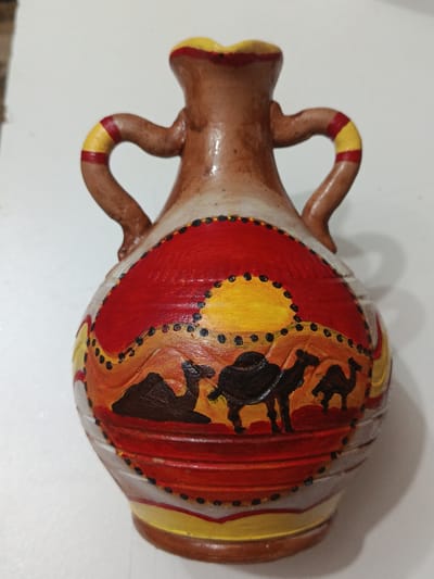 The image shows a decorative ceramic vase with a desert scene painted on it. The vase has a brown, reddish-brown, and cream color scheme. It features two handles that are decorated with red and yellow stripes. The painted scene on the body of the vase depicts a desert landscape with three camels silhouetted against a large yellow sun. The background of the scene transitions from red to orange and yellow, with dark dots creating a decorative border. The vase is placed on a white surface, likely a table or floor, and is illuminated from above, casting a shadow to the right.
Based on the provided image and context, it is not possible to determine the location of this vase within the city of Ashgabat, Turkmenistan. The image focuses solely on the object and its immediate surroundings.