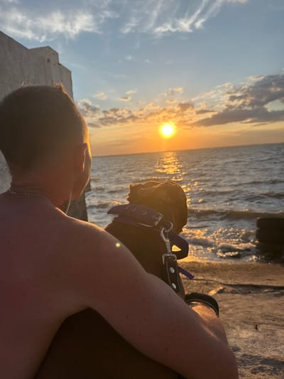 A man and his dog sit on a concrete structure facing the sea at sunset. The man is shirtless and has a chain necklace. The dog is wearing a blue collar. The sun is setting over the water, casting a golden glow on the clouds and the sea. The location is the city of Zatoka, Ukraine.