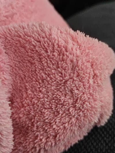 The image shows a close-up, overhead view of a fluffy pink fabric, likely a towel or blanket. The texture is soft and plush, with many fine fibers creating a fuzzy surface. The fabric is gently creased, suggesting it is being held or has been folded. The background is dark and out of focus, possibly a piece of furniture or clothing. There are no people, actions, or interactions visible in the image. The location in Bugulma, Russia, is not discernible from the visual information provided.
