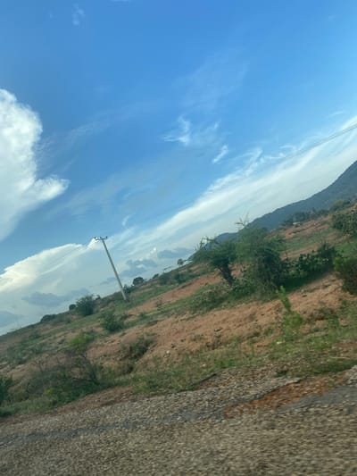 An outdoor, tilted, medium-shot captures a cloudy, bright, blue sky with a hint of a mountain range in the distance. The foreground is dominated by a rough, gray, and brown patch of road, likely gravel or pavement, with some darker material mixed in. To the left and center of the frame, a tall, wooden utility pole stands at an angle, supporting multiple power lines that stretch across the frame. The midground features a sloping, dry, reddish-brown terrain dotted with sparse green bushes and trees. The terrain appears somewhat arid and uneven, with visible signs of erosion. Towards the background, more trees and vegetation can be seen, with the mountain range serving as a backdrop under the expansive sky. The overall impression is of a rural or semi-rural landscape with evidence of infrastructure. The image does not contain any people or animals. It is not possible to determine the specific location within Gwol, Nigeria based solely on this image.