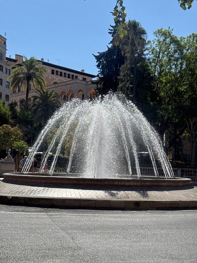 The image captures a wide shot of a large, circular fountain actively spraying water upwards and outwards in a dome-like formation. The water droplets catch the sunlight, creating a shimmering effect. The fountain is situated in a paved area with patterned tiles, surrounded by a stone rim. In the background, several people are visible standing near the fountain. Behind them, a multi-story building with arched windows and a palm tree suggest an urban setting. The sky is clear and blue, indicating a sunny day. The overall scene depicts a public space with a decorative water feature. The context of Palma, Spain is identified.