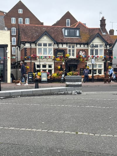 The image shows a street scene in Poole, United Kingdom, with a pub named "Jolly Sailor" prominently featured. A man in a dark jacket and light shorts is walking a brown dog on a leash on the pavement. Several other people and dogs are visible in the background, with outdoor seating areas in front of the pub. The architecture suggests a coastal town setting, with red-tiled roofs and brick buildings. A large grey concrete barrier runs across the foreground, separating the pavement from a road marked with dashed lines. The sky is overcast.