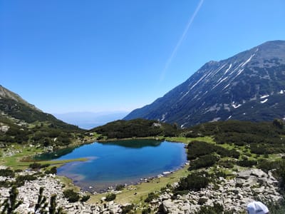 A panoramic view captures a serene mountain landscape under a clear blue sky. A tranquil, deep blue lake is the focal point, reflecting the surrounding scenery. Rocky shores dotted with green vegetation frame the water. In the background, imposing mountains rise, their slopes partially covered with snow and sparse dark green foliage, indicating a high altitude. The sky is a vibrant, unblemished blue, with a faint contrail streaking across it. The lighting suggests it is daytime, likely late morning or early afternoon, with bright sunshine illuminating the scene. The overall impression is one of natural beauty and solitude, evoking a sense of peace and the majesty of the wilderness. A small patch of lighter blue sky and distant, hazy mountains can be seen in the far background, suggesting a vast, expansive vista. In the foreground, a person wearing a white cap is partially visible, looking out at the scene, adding a human element to the grand natural setting.