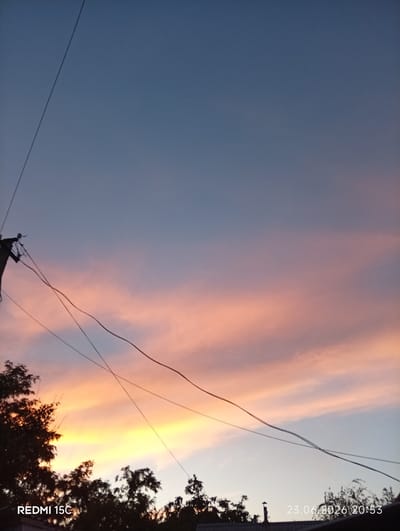 An outdoor, low-angle, full shot captures a twilight sky in Mykolaivka, Ukraine. The sky transitions from a deep blue at the top to shades of pink, orange, and yellow towards the horizon. Wispy clouds are illuminated by the setting sun, creating a vibrant, streaky effect. In the foreground, silhouetted against the bright sky, are the tops of trees and power lines. A utility pole with multiple insulators and wires is visible on the left. The timestamp "23.06.2026 20:53" and "REDMI 15C" are watermarked on the bottom right and bottom left corners respectively.