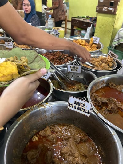 In a bustling Indonesian food stall in Cirebon, a person's arm is visible in the foreground, holding a plate of food. The plate contains rice, a yellow piece of food, and several pieces of meat with a sauce. The background shows a variety of food dishes in metal containers, including what appears to be beef rendang, semur hati & ampela (stewed liver and gizzard), pare-isi oncom (bitter melon with fermented soybean), bakwan jagung (corn fritters), and perkedel kentang (potato fritters). A woman wearing a blue hijab is visible in the middle ground, looking towards the food. The stall is brightly lit, with green walls and wooden furniture. The atmosphere is lively and suggestive of a popular eatery.