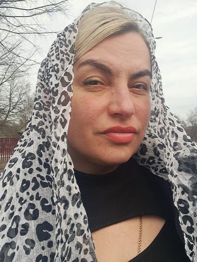 A close-up outdoor portrait shows an adult female subject in Avdot’ino, Russia. The woman has fair skin, blonde hair visible beneath a white, translucent headscarf with a black leopard print pattern. Her dark eyebrows are visible, and her lips are colored with reddish-orange lipstick. She wears a black garment, possibly a top or jacket, which includes a partial cutout revealing a gold chain on her chest. Her gaze is directed towards the camera. The background, largely out of focus, features bare tree branches against an overcast, light gray sky. A segment of a reddish-brown fence or structure is visible behind her left shoulder.