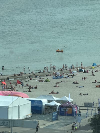 This is a high-angle, slightly tilted shot of a crowded beach in Gdynia, Poland. The scene is dominated by the light gray sand and the rippling, light blue water. A multitude of people are spread across the beach, engaged in various activities. Some are sunbathing on towels or directly on the sand, while others are standing, walking, or interacting with each other. The water is calm, with a few small yellow buoys visible in the distance. One person is on a bright orange inflatable raft in the water.

Further back from the water, there are several tents and structures, suggesting an event or a vendor area. Some of these tents are white and large, providing shade. One prominent tent has purple and blue branding with some text. A darker, blue tent is also visible. There are also metal scaffolding structures, possibly part of the event setup.

The time of day appears to be daytime, likely late morning or afternoon, given the bright sunlight casting shadows. The weather seems pleasant, conducive to beach activities. The atmosphere appears lively and recreational, with a large gathering of people enjoying a summer day.

Visible text on the purple and blue tent includes "ZEGNI PUZO" and potentially other Polish words or branding. There is also text on a sign above the tents.