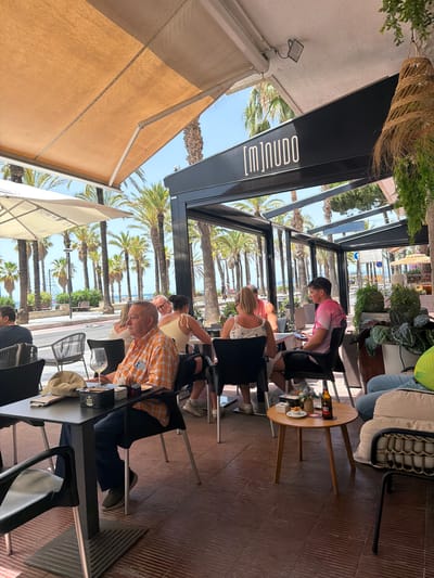 This media file depicts a sunny day at an outdoor cafe in Salou, Spain, with palm trees and a beach visible in the background. The scene is viewed from a low angle, looking upwards towards the cafe's awnings and structure. Several people are seated at tables, engaged in various activities.

In the foreground, a man in an orange and white checkered shirt is seated at a table, looking down. A glass of white wine and a hat are on the table in front of him. To his left, another person is partially visible. Further back, several other individuals are seated at tables, some appearing to be engaged in conversation or using their phones. One person is wearing a pink shirt and looking at a mobile device.

The cafe has a modern, open-air design with dark metal framing and wooden flooring. A prominent black awning extends over the seating area, displaying the white text "[m]inudo" in a distinctive font. The overall atmosphere appears relaxed and casual, indicative of a typical day at a seaside establishment. The bright sunlight suggests it is daytime, likely late morning or afternoon. The presence of palm trees and the general ambiance strongly point to a Mediterranean coastal location.