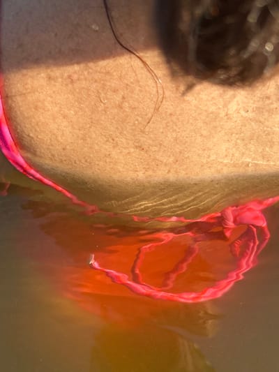 The image shows a close-up of a person's shoulder and neck, partially submerged in water. A pink bikini strap is visible around the shoulder. The water distorts the view of the submerged bikini, creating an orange and red hue. The background includes skin texture with water droplets and a shadow cast by an unseen object, possibly hair or a piece of clothing. The location in Pribrezhnyi, Russia, is not evident from the visual information provided in the image.