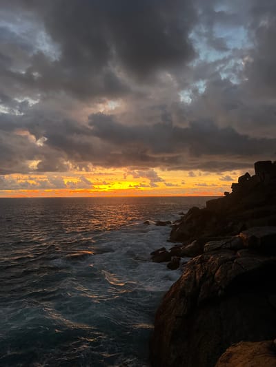 This is a vivid landscape photograph capturing a sunset over the ocean, viewed from a rocky coastline, likely in Rumassala, Sri Lanka. The image is characterized by dramatic lighting and a striking contrast between dark and bright elements.

The horizon is dominated by a brilliant band of orange and yellow light, indicating the sun setting or rising low over the water. This warm glow extends across the distant sea, creating shimmering reflections on the water's surface.

The sky on the left side of the frame is filled with heavy, dark grey and blue-grey clouds, suggesting a cloudy or stormy atmosphere, yet pockets of lighter, illuminated clouds are visible, catching the warm hues from the horizon.

On the right, a rugged, dark rocky cliff or headland juts into the frame, silhouetted against the bright horizon. Its texture is visible, with some surfaces catching faint golden highlights from the low sun, while shadowed areas remain deep and dark. The base of the cliff meets the ocean, where waves are visible, their ripples reflecting the ambient light.

The ocean itself appears deep blue-grey, with numerous small waves and whitecaps, particularly noticeable closer to the shore and cliff. The interplay of light and shadow on the water creates a dynamic, textured surface.

The overall impression is one of dramatic natural beauty, highlighting the raw power of the sea and sky during a colorful sunset, with the dark cliffs providing a stark, grounding contrast. There is no visible text or identifiable human activity.