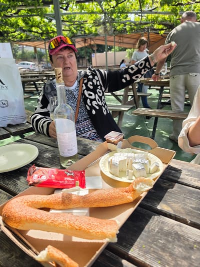 Here is a description of the image:
A woman sits at a wooden picnic table with food and drinks. She is wearing a black and white patterned sweater and a pair of brightly colored sunglasses perched on her forehead. She holds a small piece of food in her raised hand.
On the table are a bottle of wine, a platter of cheese, a package of chocolate, and twisted sesame seed bread in a cardboard container. There is a paper bag and a plate on the table, and an open bottle of wine is also visible.
The picnic table is located outdoors, under a canopy of greenery. The scene is in Mate Yehuda Regional Council, Israel, as inferred from the text on the bottle label. Other people are visible in the background, seated at similar tables.