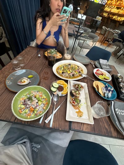 The image captures a restaurant setting with a woman taking a photo of various dishes on a wooden table. The location appears to be in the city of Santiago de Querétaro, Mexico, as inferred from the restaurant's name "Santo Mar" visible on a napkin.

The woman is seated at the table, holding a smartphone with a distinctive case and taking a picture of the assortment of food. She is wearing a blue top and a silver bracelet. 

Several plates of food are arranged on the table, including a colorful salad with avocado and other ingredients in a green bowl, a ceviche-style dish in a white bowl, some taco-like snacks in a blue bowl, and other small dishes. There's also a glass of liquid, possibly a cocktail, and silverware placed alongside the plates.

The restaurant's ambiance is suggested by the background, which shows a bar area with lighting and other seating. The setting appears to be an upscale dining establishment.