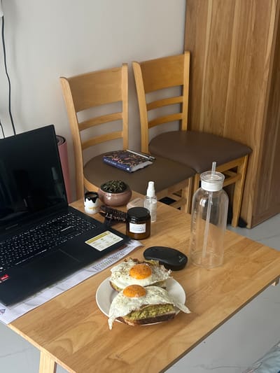A slightly elevated, angled shot focuses on a wooden desk with breakfast, work, and personal items, likely indoors. On the desk is an open laptop with a black screen and visible keyboard, alongside a plate of food featuring fried eggs on toast. A black computer mouse rests near the plate. Other objects present on the desk include a clear water bottle, a small potted plant, a hair clip, a small decorative figure, and a cylindrical jar with text in a Cyrillic script. There is also a book placed between the desk and a wooden chair. Behind the chair, a pink yoga mat is partially visible. The image indicates a personal or work space. The location in the city of Nam Natural Heritage Area Trang, Vietnam, is not evident from the image, as there are no recognizable external environmental cues.