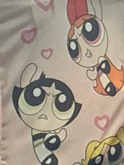 The image is a close-up of a pink textile with a pattern of cartoon characters and hearts. Two of the characters are clearly visible: one with black hair and a star on her forehead, and another with reddish-orange hair and a bow. Both characters have large, round eyes. A third character's blond hair and eye are partially visible at the bottom right. The hearts are small and pink. The textile appears to be fabric, possibly a blanket or clothing. There is no discernible location in Enevo, Bulgaria.